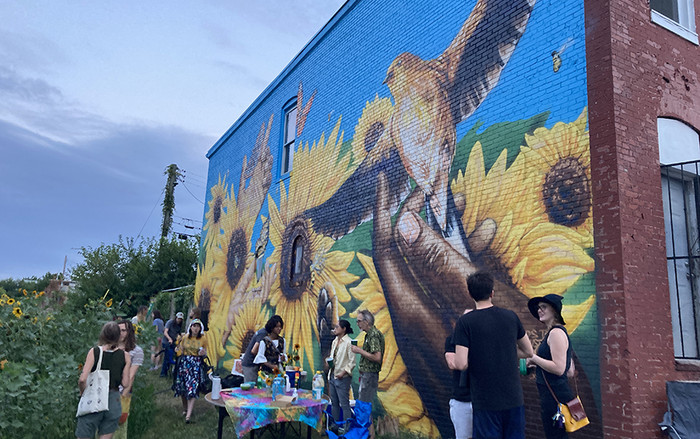 people outside in a sunflower garden in a city with a mural with a brown hand releasing a bird